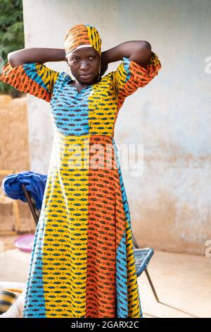 In questa immagine, una giovane casalinga nera alta che indossa un vestito lungo africano tradizionale colorato sta fissando il suo copricapo corrispondente Foto Stock