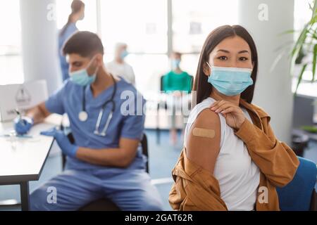 Dopo il concetto di vaccinazione. Ritratto di vaccinato Asian Woman in Mask che mostra braccio ottenere iniezione antivirale, giovane Signora seduta con Rolled Up Sleev Foto Stock