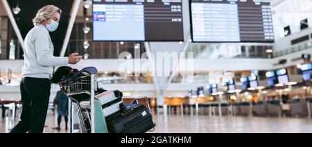 Uomo che indossa una maschera a piedi con il trolley bagagli al terminal dell'aeroporto. Viaggiatore maschile che usa il telefono cellulare all'aeroporto. Foto Stock