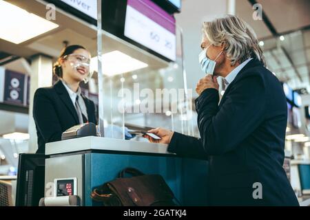 Uomo d'affari in viaggio che effettua il check-in in aeroporto durante la pandemia di COVID-19. Passeggero maschile in maschera e sicurezza aeroporto dietro acrilico Foto Stock