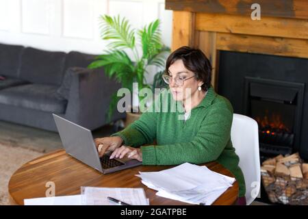 Foto di una donna anziana sta riempiendo le forme che si siedono davanti al monitor del laptop nell'interno domestico. Lavora online e utilizza tutorial per il riempimento Foto Stock