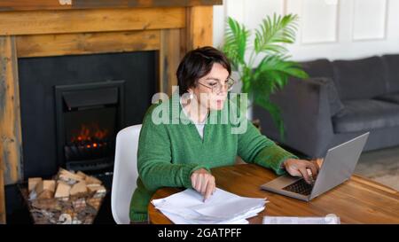 La donna anziana sta riempiendo le forme che siedono davanti al monitor del laptop nell'interno domestico. Lavora online e utilizza tutorial per riempire la sua tassa per Foto Stock