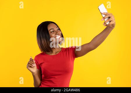 Happy Black femmina con smartphone rendendo Selfie Ritratto in posa e avendo divertente in piedi su sfondo giallo Studio. Persone e Gadget, Social Media Foto Stock