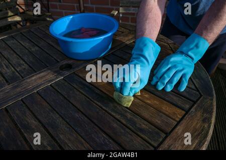 L'uomo con i guanti di gomma blu sta pulendo un tavolo da giardino di legno scuro all'inizio della primavera, spazio di copia, fuoco selezionato Foto Stock