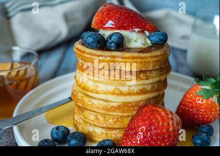 Una pila di frittelle con miele, fragola, bacca blu e panna a frusta sul piatto del tavolo bianco, delizioso dessert per colazione. Foto Stock
