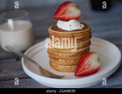 Vista chiara della pila di frittelle con bastone di miele, fragola, bacca blu e panna frusta in cima nel piatto, delizioso dessert con latte per la colazione. Foto Stock