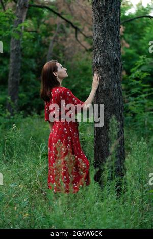 Salvare e piantare alberi, salvare la Terra e la natura, stare per gli alberi, salvare le foreste e guarire il clima. Donna in un vestito rosso che cammina nella foresta, abbracciando gli alberi Foto Stock