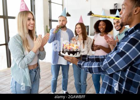 Amici diversi che danno la torta di compleanno yummy alla signora blonde eccitata, avendo festa a casa. Happy millennial indossando cappelli festosi, con la mano che si aggrappano Foto Stock