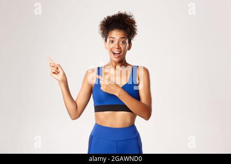 Sana e attraente sportivo in outfit blu sport, puntando le dita sinistra e guardando sorpreso, mostrando marchio o logo, sfondo bianco Foto Stock
