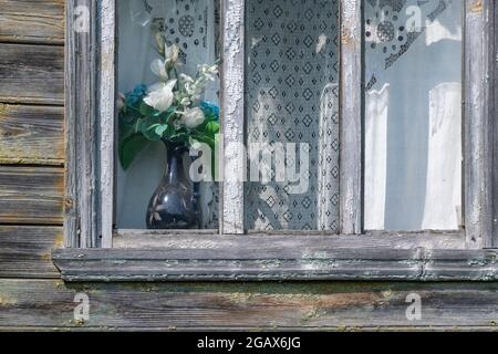 Finestra in legno sbucciato invecchiato decorata con tende bianche tradizionali e un bouquet di fiori sintetici in un vaso in ceramica dipinta a mano blu Foto Stock