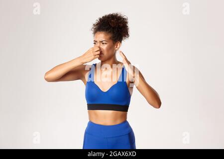 Studio shot di atleta afro-americana disgustata che lamenta cattivo odore, naso chiuso e scuotere la mano da un terribile pupore, in piedi contro bianco Foto Stock