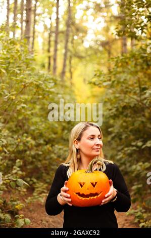 Primo piano foto di giovane bionda femmina che tiene spaventosa zucca artigianale dal viso, in piedi isolato sullo sfondo della foresta autunnale. Vacanze autunnali, hallo Foto Stock