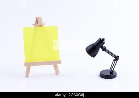 Easel in legno in miniatura con tela quadrata colorata vuota o carta Memo - Mockup. Mini supporto in legno con tavola da disegno pulita e piccola lampada da tavolo nera su sfondo bianco, Copy Space Foto Stock