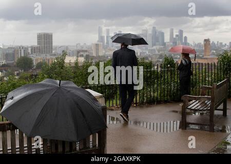 30/07/2021. Londra, Regno Unito. I membri del riparo pubblico sotto gli ombrelloni mentre guardano fuori su una vista di Canary Wharf durante una tempesta di pioggia su Point Hill Foto Stock