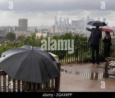 30/07/2021. Londra, Regno Unito. I membri del riparo pubblico sotto gli ombrelloni mentre guardano fuori su una vista di Canary Wharf durante una tempesta di pioggia su Point Hill Foto Stock