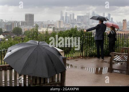 30/07/2021. Londra, Regno Unito. I membri del riparo pubblico sotto gli ombrelloni mentre guardano fuori su una vista di Canary Wharf durante una tempesta di pioggia su Point Hill Foto Stock