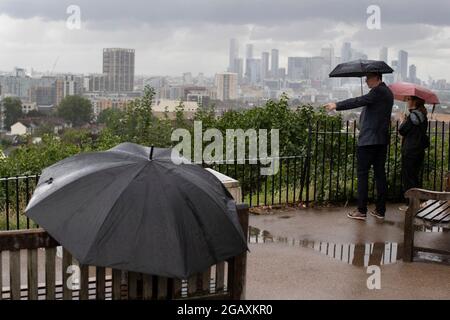 30/07/2021. Londra, Regno Unito. I membri del riparo pubblico sotto gli ombrelloni mentre guardano fuori su una vista di Canary Wharf durante una tempesta di pioggia su Point Hill Foto Stock