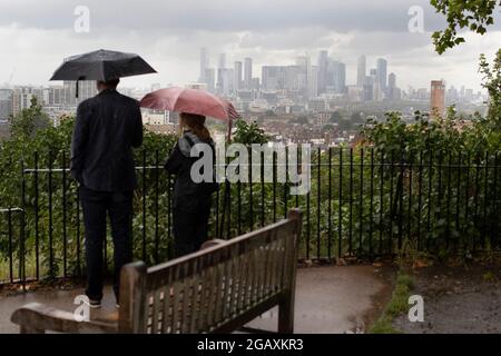 30/07/2021. Londra, Regno Unito. I membri del riparo pubblico sotto gli ombrelloni mentre guardano fuori su una vista di Canary Wharf durante una tempesta di pioggia su Point Hill Foto Stock