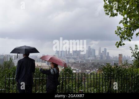 30/07/2021. Londra, Regno Unito. I membri del riparo pubblico sotto gli ombrelloni mentre guardano fuori su una vista di Canary Wharf durante una tempesta di pioggia su Point Hill Foto Stock