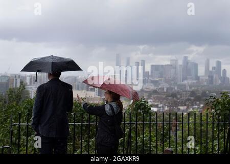 30/07/2021. Londra, Regno Unito. I membri del riparo pubblico sotto gli ombrelloni mentre guardano fuori su una vista di Canary Wharf durante una tempesta di pioggia su Point Hill Foto Stock