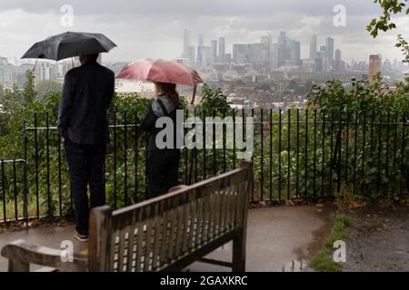 30/07/2021. Londra, Regno Unito. I membri del riparo pubblico sotto gli ombrelloni mentre guardano fuori su una vista di Canary Wharf durante una tempesta di pioggia su Point Hill Foto Stock