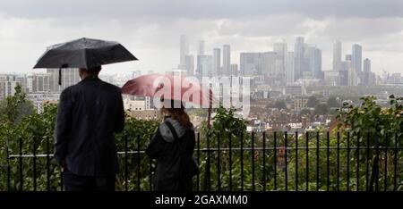 30/07/2021. Londra, Regno Unito. I membri del riparo pubblico sotto gli ombrelloni mentre guardano fuori su una vista di Canary Wharf durante una tempesta di pioggia su Point Hill Foto Stock