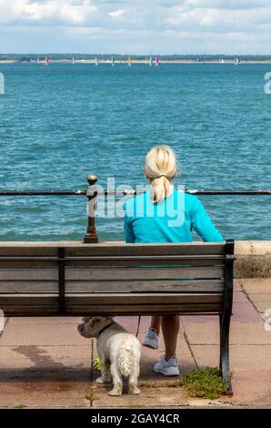 signora con cane su panca di legno, signora con cane seduta, cane da passeggio femminile al mare, femmina seduta su un sedile con cane piccolo, femmina con cane piccolo. Foto Stock