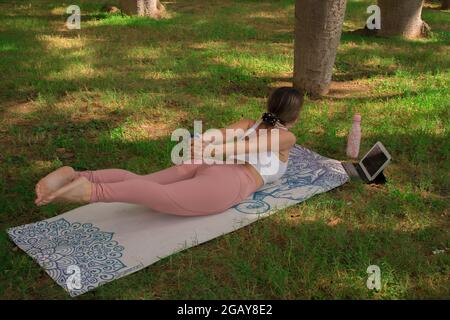 Giovane donna che fa la posa del grasshopper mentre è distendersi all'aperto nel parco della città in una giornata soleggiata in estate. Pilates Outdoor 2021. Foto Stock