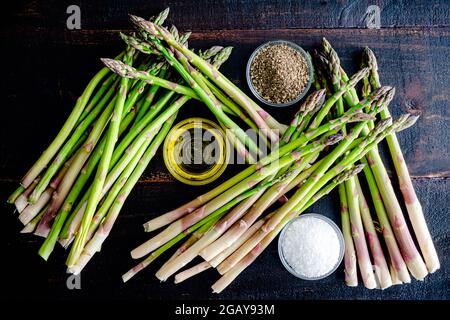 Asparagi arrosto ingredienti su un legno scuro sfondo: Spighe di asparagi crudi, olio d'oliva, sale e pepe su un tavolo di legno Foto Stock