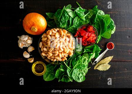 Fagioli spagnoli vegani con pomodori ingredienti su un tavolo di legno: Fagioli cannellini crudi, pomodori a dadini, foglie di spinaci e spezie per preparare un piatto vegano Foto Stock