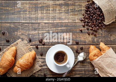 Tazza di caffè, fagioli arrostiti, croissant su fondo di legno. Concetto di colazione con spazio fotocopie. Vista dall'alto, disposizione piatta. Foto Stock