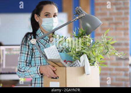 Donna in maschera medica protettiva si alza con scatola e oggetti personali in ufficio Foto Stock