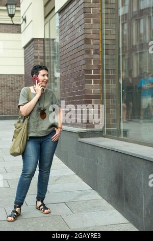 Bella donna di mezza età con un telefono in città. La donna sta parlando emotivamente al telefono. Foto Stock