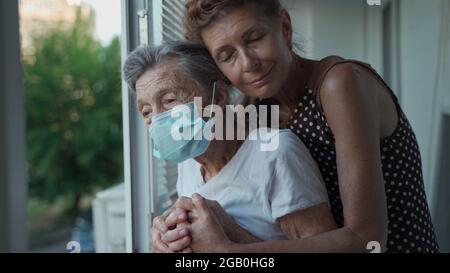 Donna solitaria anziana di 90 anni in maschera medica e sua figlia matura in piedi in abbraccio guardando fuori finestra in casa di cura. Visita di una figlia adulta Foto Stock