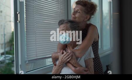 Figlia adulta abbracciando con amorevole madre anziana 90 anni in maschera in piedi alla finestra, parenti di diverse generazioni, donne in piedi Foto Stock