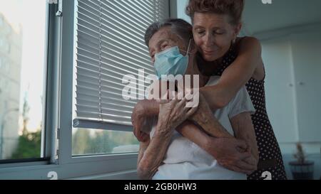 Figlia adulta abbracciando con amorevole madre anziana 90 anni in maschera in piedi alla finestra, parenti di diverse generazioni, donne in piedi Foto Stock