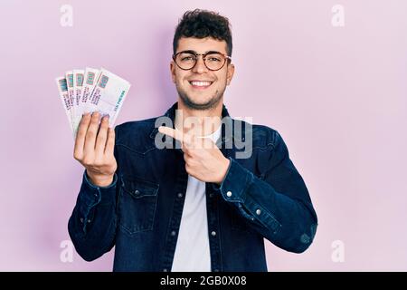 Giovane uomo ispanico che tiene banconote in sterline egiziane sorridendo felice indicando con mano e dito Foto Stock