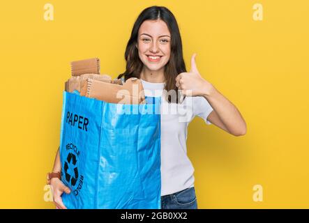 Giovane bella donna che tiene riciclaggio wastbasket con carta e cartone sorridente felice e positivo, pollice su facendo eccellente e segno di approvazione Foto Stock