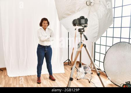 Donna ispanica di mezza età che si pone come modello in studio di fotografia scettica e nervosa, disapprovando l'espressione sul viso con le braccia incrociate. Perso negativo Foto Stock