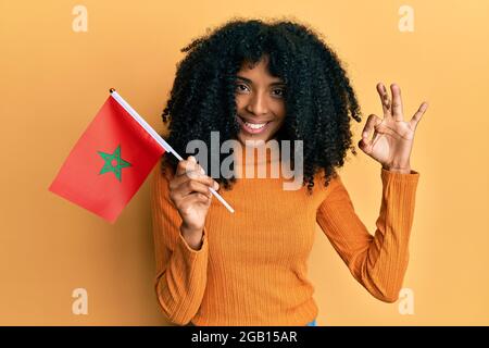 Donna afroamericana con i capelli afro tenendo bandiera marocco facendo ok segno con le dita, sorridente amichevole gesturing eccellente simbolo Foto Stock