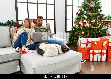 Giovane coppia che usa il laptop seduto da albero di natale facendo gesto italiano con mano e dita espressione sicura Foto Stock
