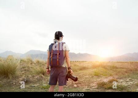 vista posteriore di un fotografo asiatico che guarda la vista alla luce del sole del mattino con la fotocamera in mano Foto Stock