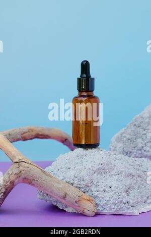 Vasetto di vetro ambrato di liquido cosmetico con contagocce su pietra e legno, siero di faccia o acido ialuronico per la cura della pelle del viso su sfondo blu in una e naturale Foto Stock