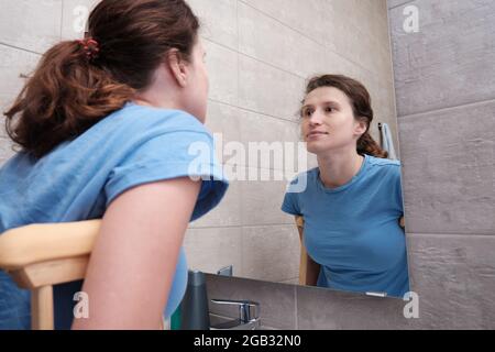 Una ragazza sulle stampelle si guarda nello specchio del bagno. Riabilitazione dopo una frattura. Foto Stock
