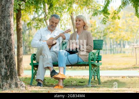 Felice coppia di amore anziano in romanticismo seduto e suonare Ukulele strumento musicale sulla sedia nel parco Foto Stock