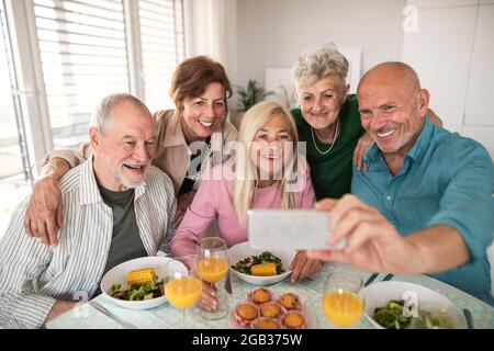 Gruppo di amici anziani che hanno festa all'interno, prendendo selfie quando si mangia al tavolo. Foto Stock
