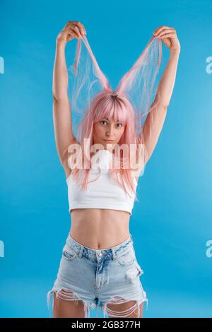 Divertente modello femminile in abbigliamento casual giocando con capelli rosa, tenendo le trefoli di esso sulla sua testa, in piedi isolato su sfondo blu studio Foto Stock