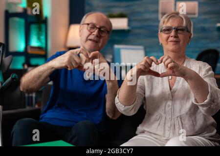 Famiglia anziana romantica che crea il simbolo della forma del cuore utilizzando le mani per la fotocamera a casa. Vecchio marito sposato e moglie seduti insieme sul divano facendo amore concetto gesto gesto sentimento allegro Foto Stock