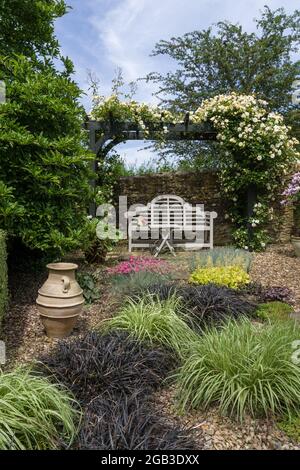 Un giardino inglese in estate, Gayton, Northamptonshire, UK; arbour, panca bianca e piante che crescono in ghiaia. Foto Stock