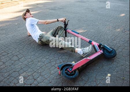 Un giovane è entrato in un incidente su uno scooter elettronico, si trova sull'asfalto e grida di dolore. Foto Stock